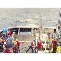 A pressure-compensated low-frequency source is deployed by Scripps Institution of Oceanography personnel off the USS Safeguard (ARS-50), circa 1986. Note the circular source in the standing equipment cage. It is pressure compensated by the four large compressed air flasks below it. Primary lithium cells and control electronics are in the vertical cylinders flanking the source to either side. The author is pictured on the far left. Lab mates Brian Dushaw, Steve Abbott, Doug Peckham, and Bob Trues