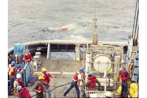 A pressure-compensated low-frequency source is deployed by Scripps Institution of Oceanography personnel off the USS Safeguard (ARS-50), circa 1986. Note the circular source in the standing equipment cage. It is pressure compensated by the four large compressed air flasks below it. Primary lithium cells and control electronics are in the vertical cylinders flanking the source to either side. The author is pictured on the far left. Lab mates Brian Dushaw, Steve Abbott, Doug Peckham, and Bob Trues