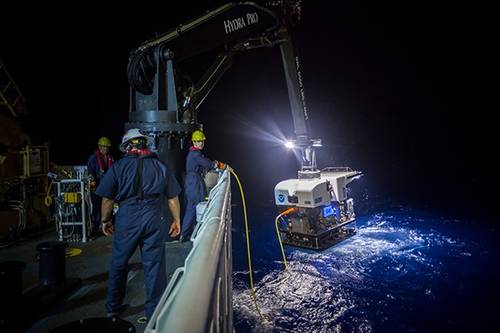 Crew members aboard a NOAA ship in 2016 use a crane to hoist a remotely operated vehicle (ROV) from the water at night. NOAA is seeking proposals for new ocean survey ships that can deploy a variety of equipment, including ROVs like the one pictured here. Credit: NOAA