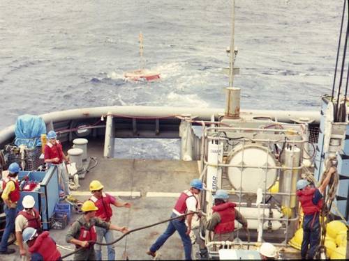 A pressure-compensated low-frequency source is deployed by Scripps Institution of Oceanography personnel off the USS Safeguard (ARS-50), circa 1986. Note the circular source in the standing equipment cage. It is pressure compensated by the four large compressed air flasks below it. Primary lithium cells and control electronics are in the vertical cylinders flanking the source to either side. The author is pictured on the far left. Lab mates Brian Dushaw, Steve Abbott, Doug Peckham, and Bob Trues