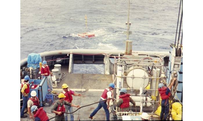 A pressure-compensated low-frequency source is deployed by Scripps Institution of Oceanography personnel off the USS Safeguard (ARS-50), circa 1986. Note the circular source in the standing equipment cage. It is pressure compensated by the four large compressed air flasks below it. Primary lithium cells and control electronics are in the vertical cylinders flanking the source to either side. The author is pictured on the far left. Lab mates Brian Dushaw, Steve Abbott, Doug Peckham, and Bob Trues